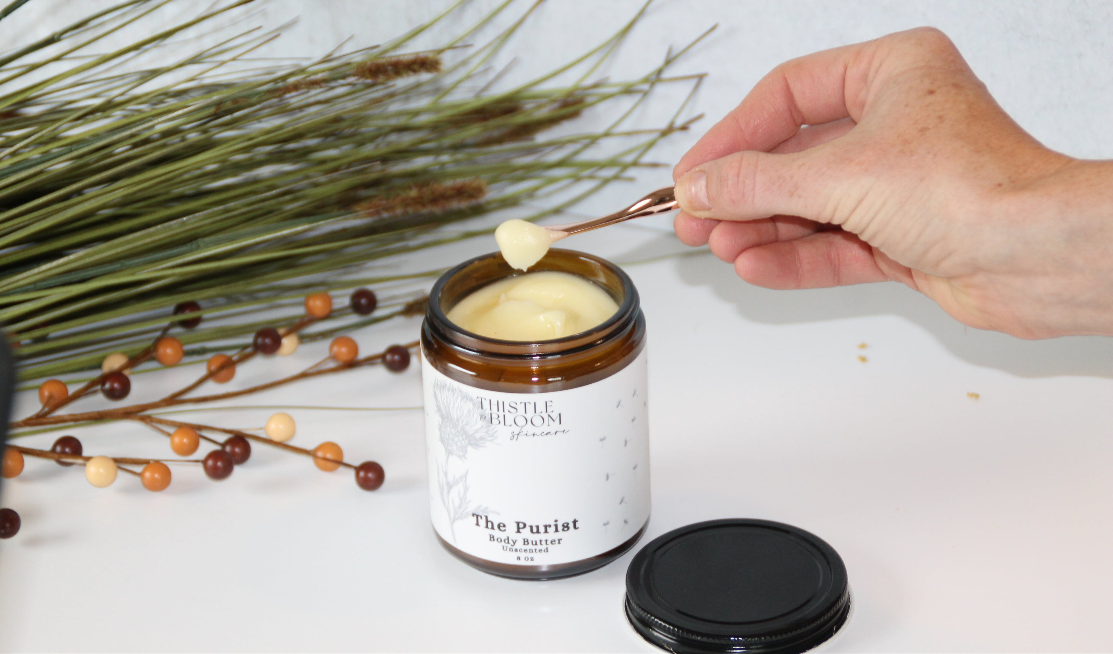 Person using a spoon to scoop out a product from a jar labeled 'The Purist' on a white surface with decorative elements.